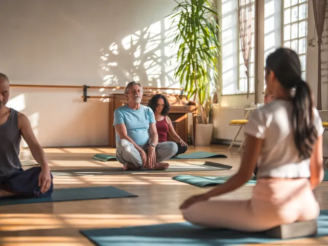 Une séance de yoga au sol dans une salle lumineuse, trois personnes face à la prof
